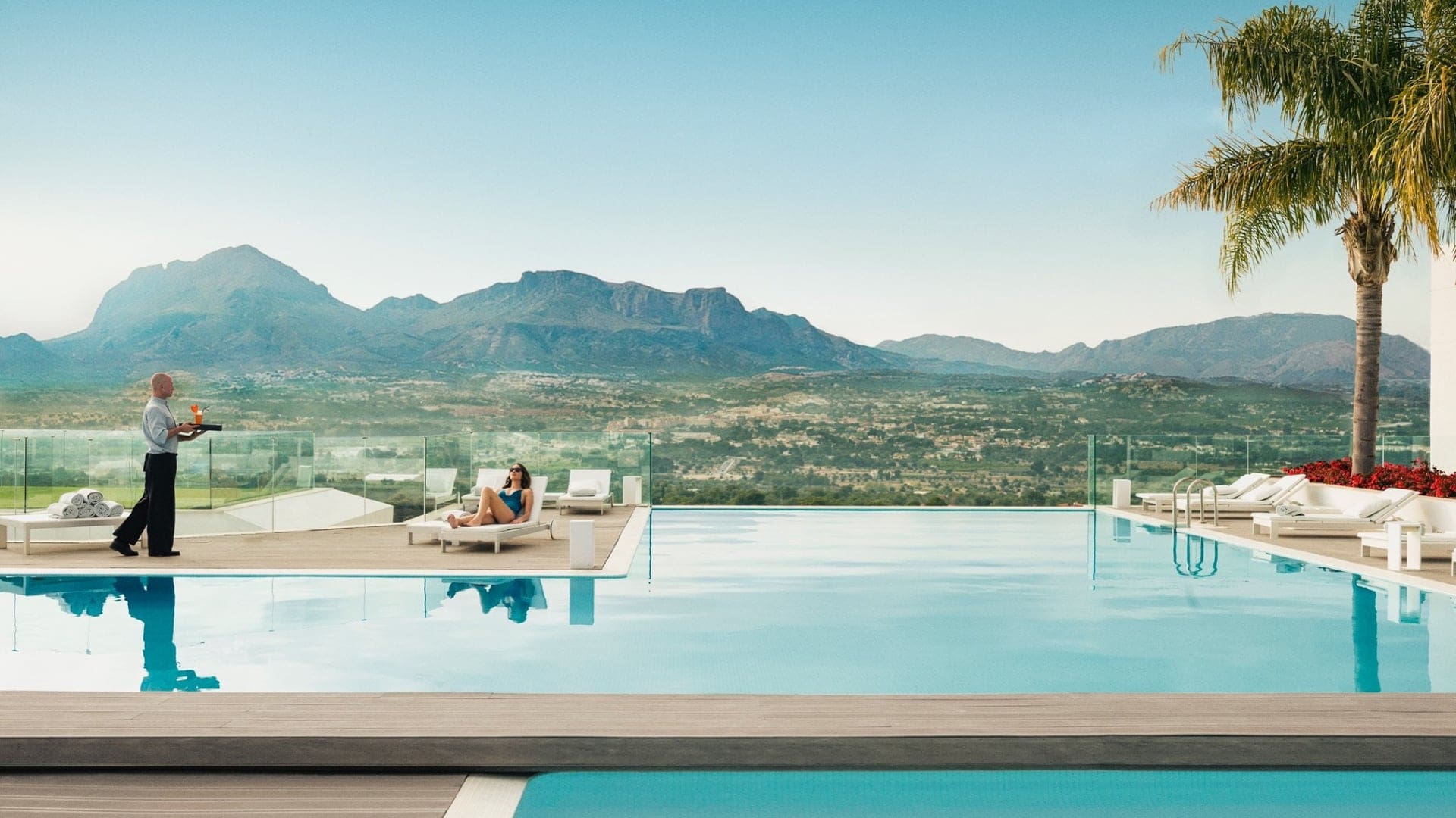 A outfoor swimming pool with a woman relaxing on a sunbed with a waitress walking towards her.