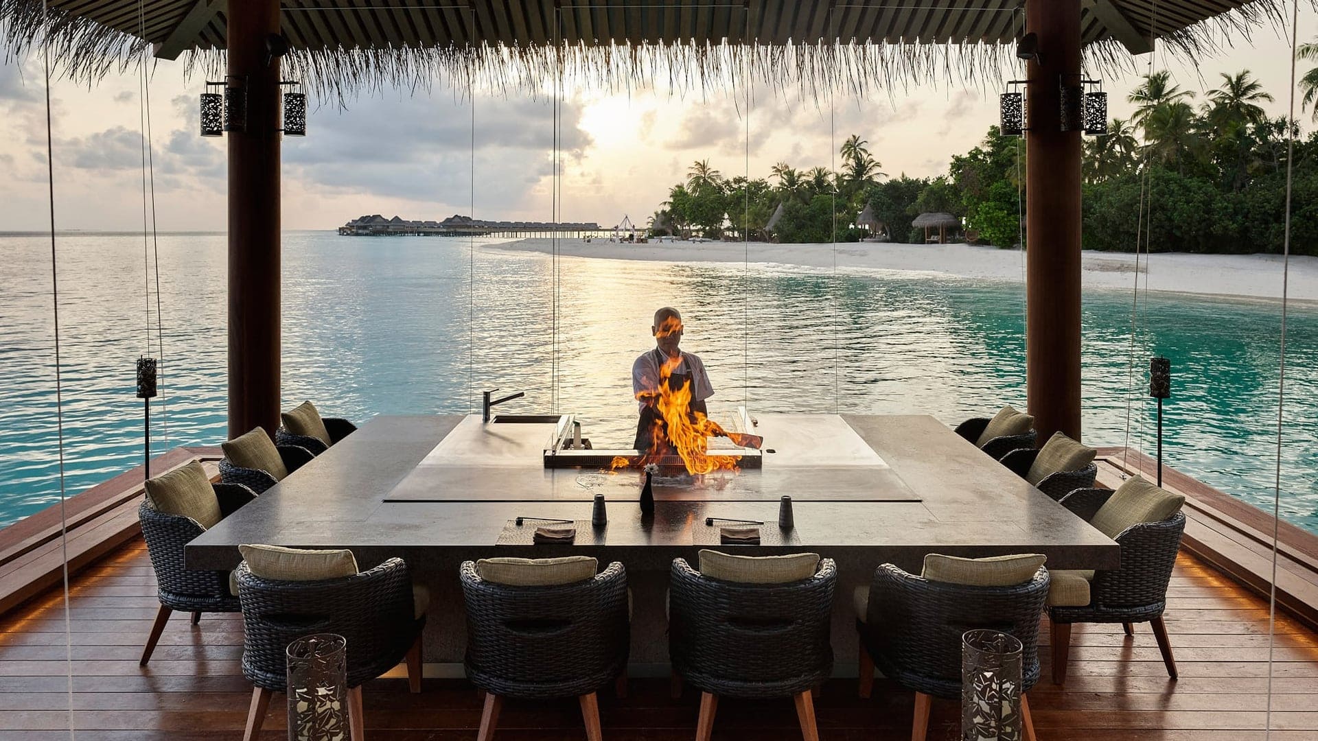 Private dining table, with fire and man in white uniform and view of JOALI Maldives island and the water, in wooden and black decor.