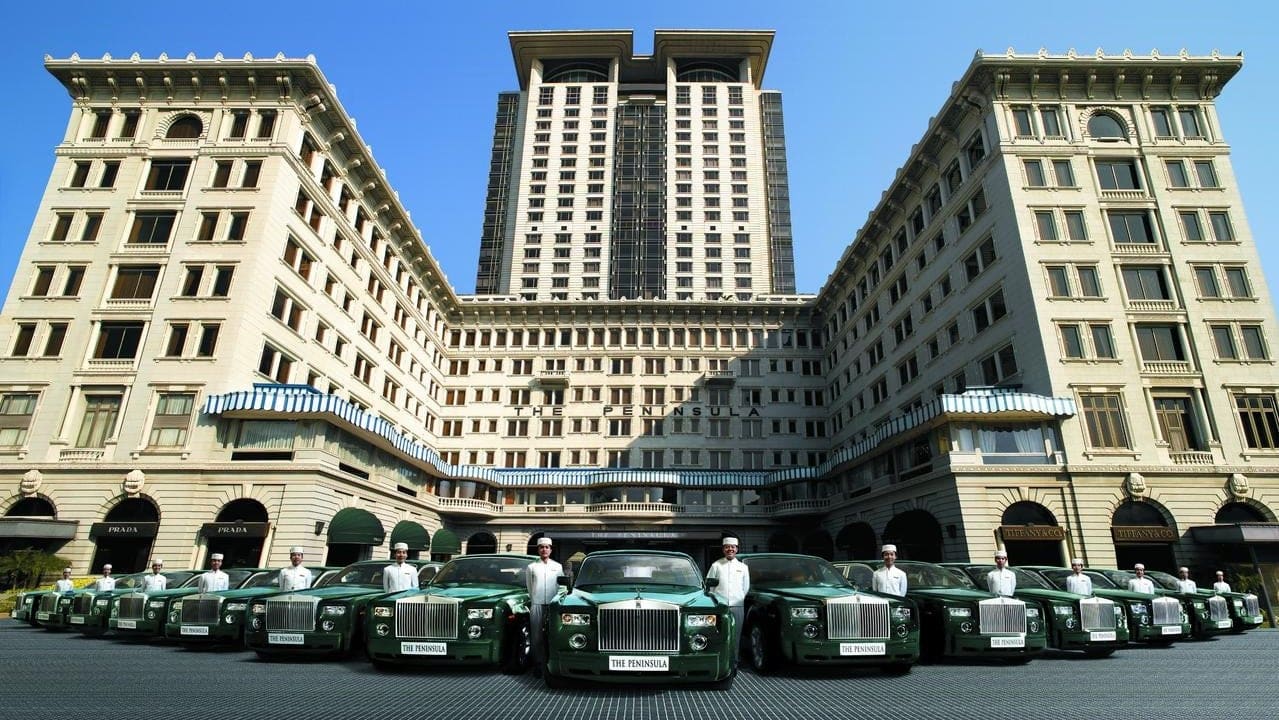 Outside view of The Peninsuala - Hong Kong with 13 dark green bentley cars parked outside and 14 hotel assistans in white beside the cars.