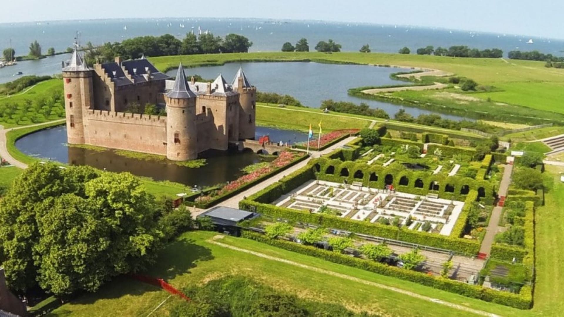 Bird perspective of Muiderslot Castle with beautiful garden, lake surrounding the castle and the castle a large brick building with roof sylinder and high fences.