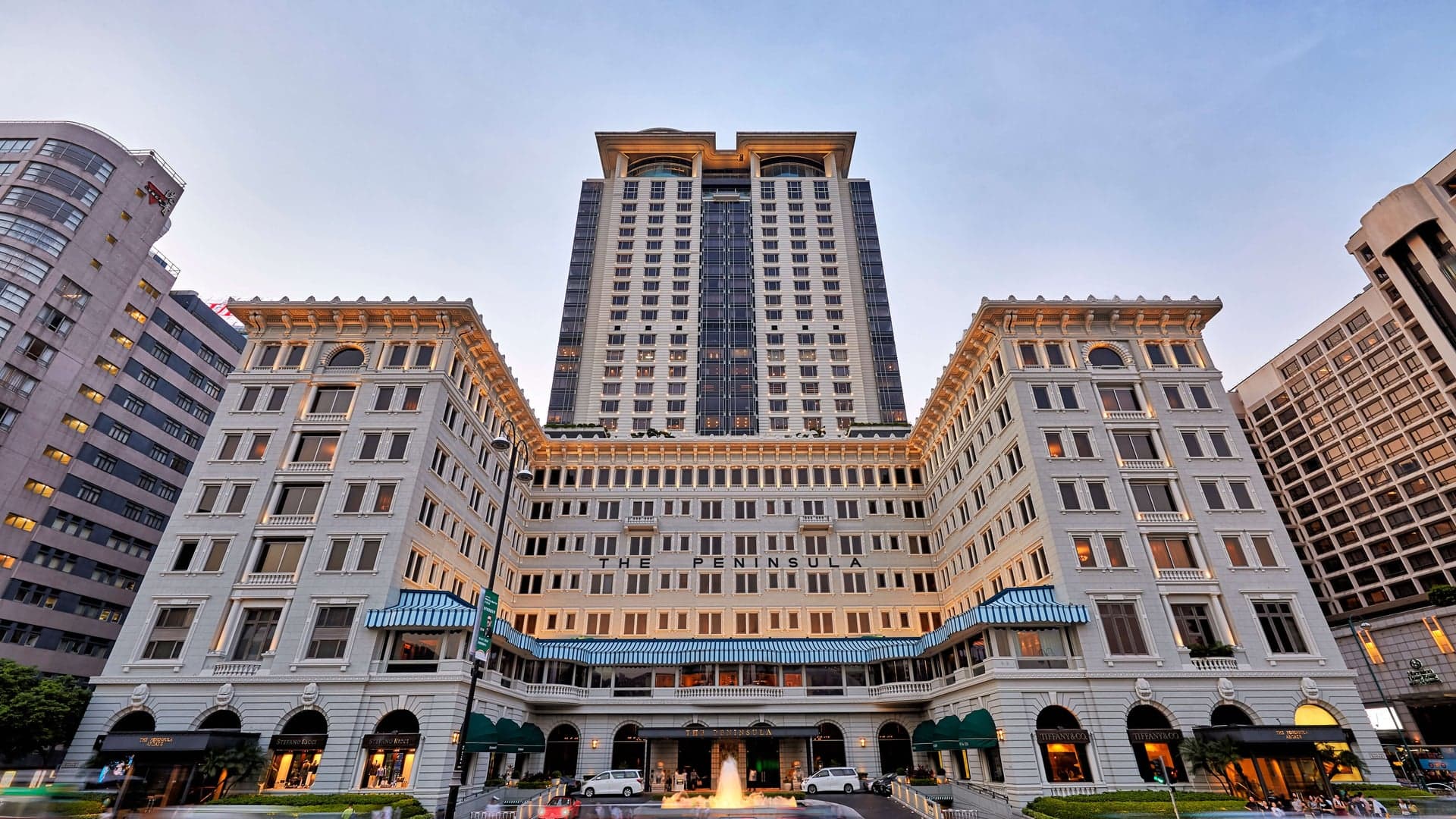 In front of The Peninsuala in Hong Kong, a large classical building with fountain and text saying: THE PENINSUALA.