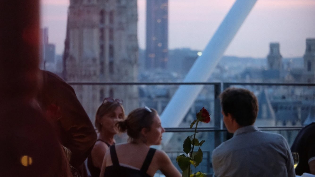 Two women in black and man in blue shirt, rose in the middle and view of Paris at Cafe Gerorges Pompiduo.