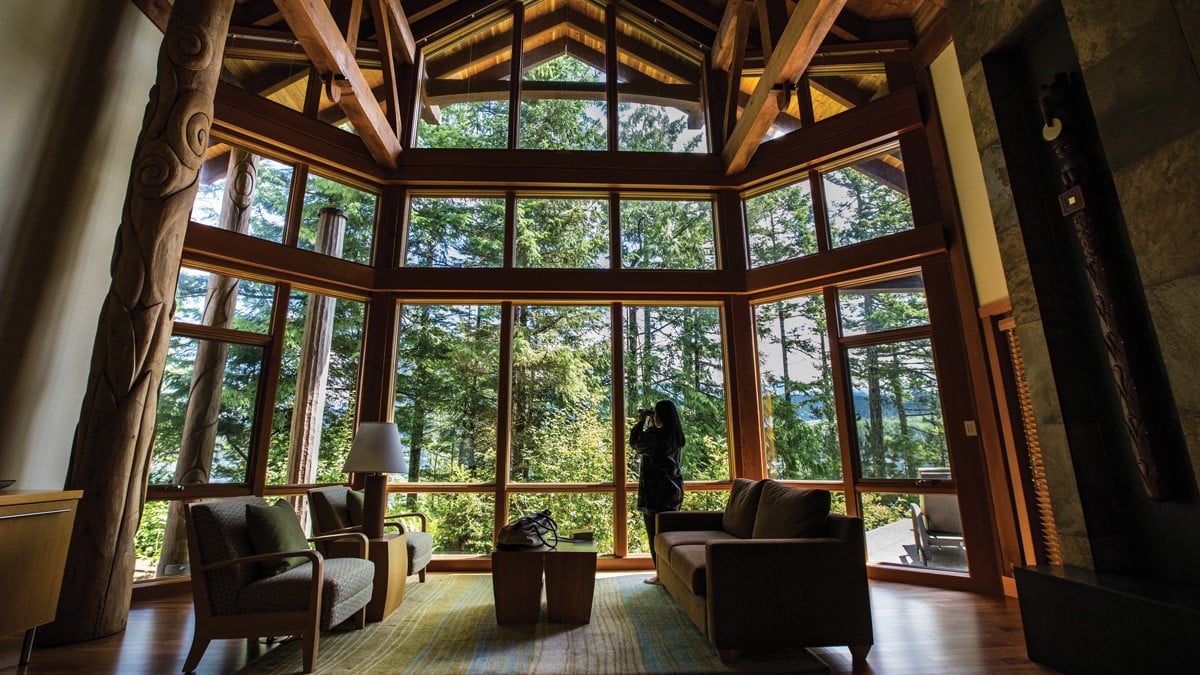 A woman looking at the woods through large wooden windows inside a cabin