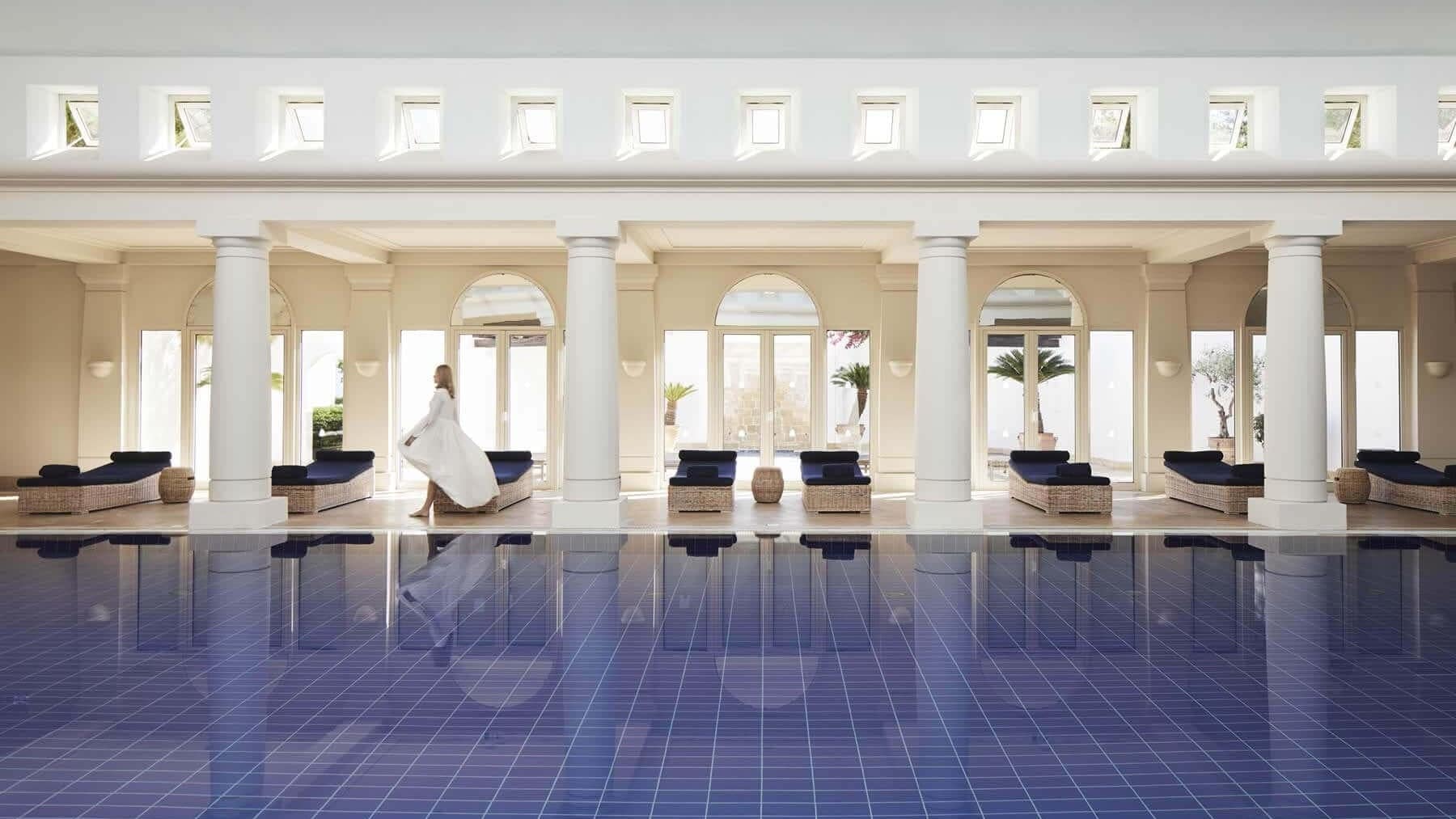 A indoor swimming pool with blue sunbeds, white columns and a woman walking by the sunbeds.