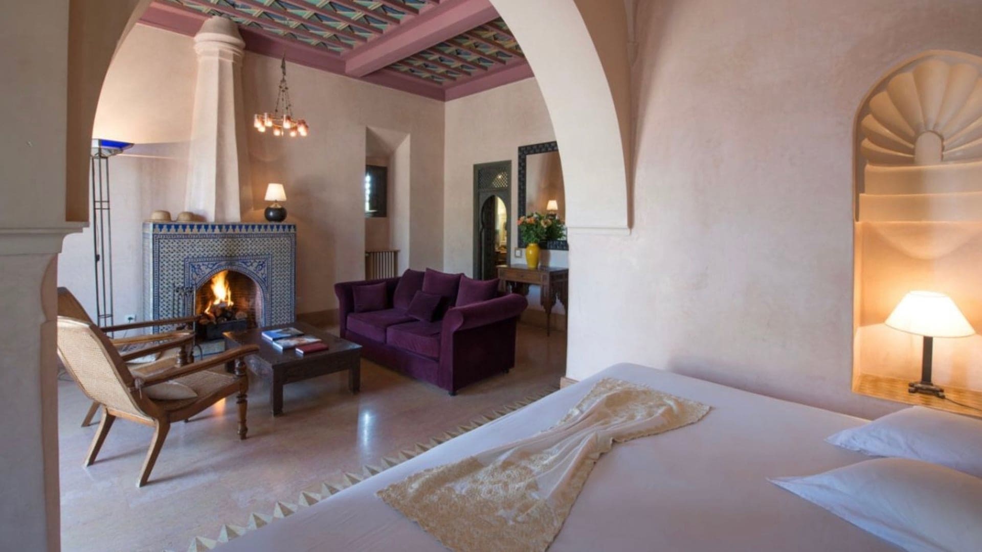White bedroom with bedside lamp and purple sofa living room area with fireplace at Les Deux Tours
