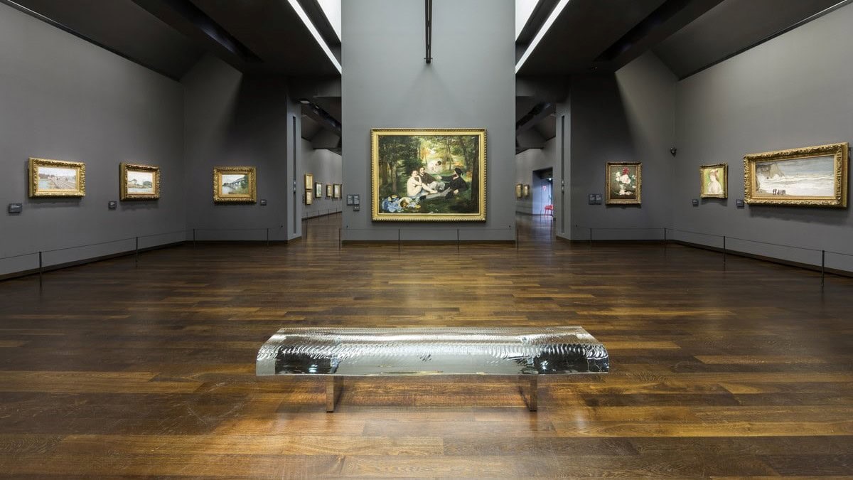 Large room with wooden floor, one chair and grey walls filled with painting at Musee D'Orsay