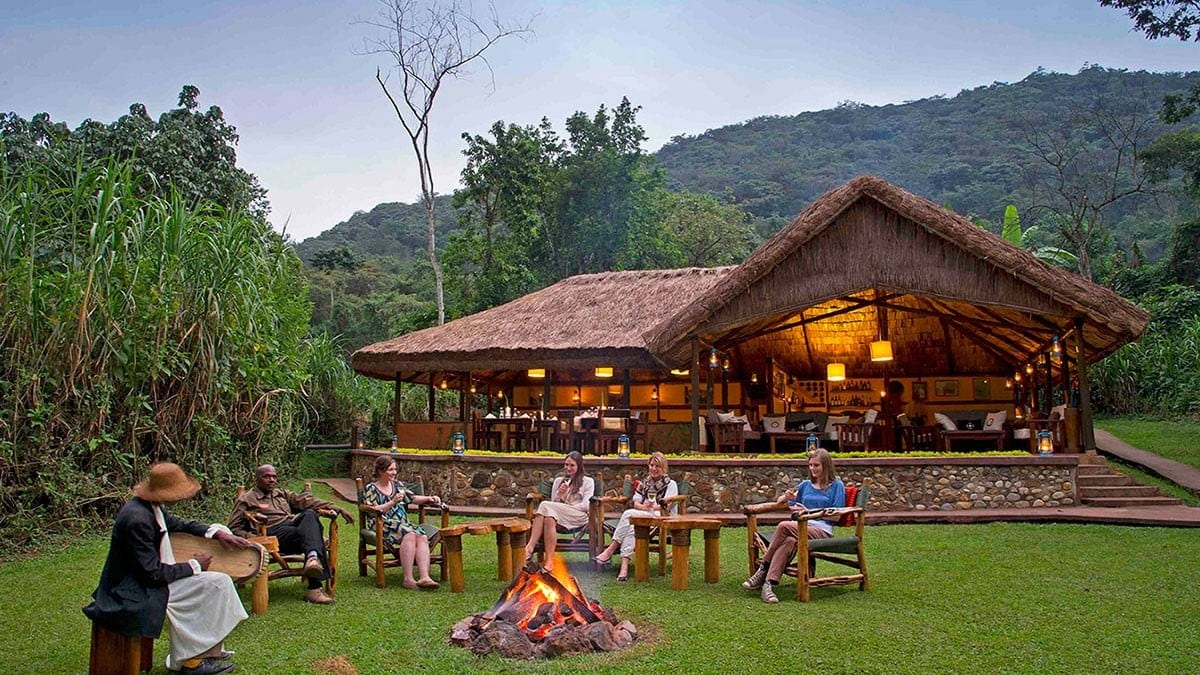 Four women and two men sitting around a campftire next to a large brown dining and bar area surrounded by nature
