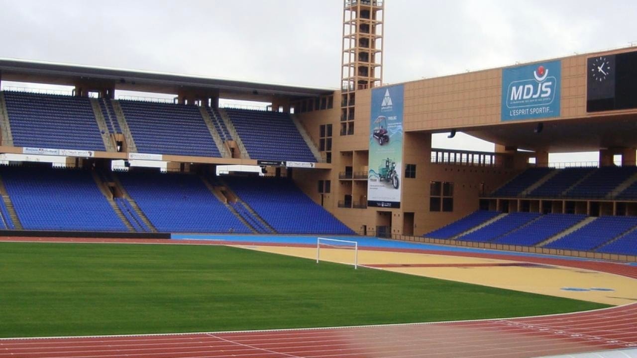 Stadium with green field and blue seats, with a red track.