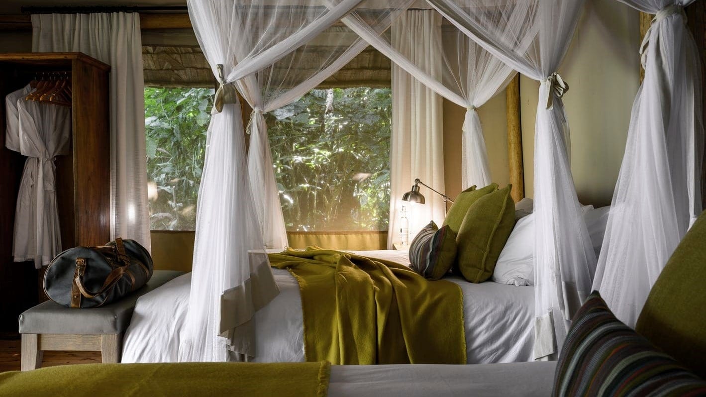 White bed with green varpet and pillows and a blue bag with a large nedside window with a view to trees