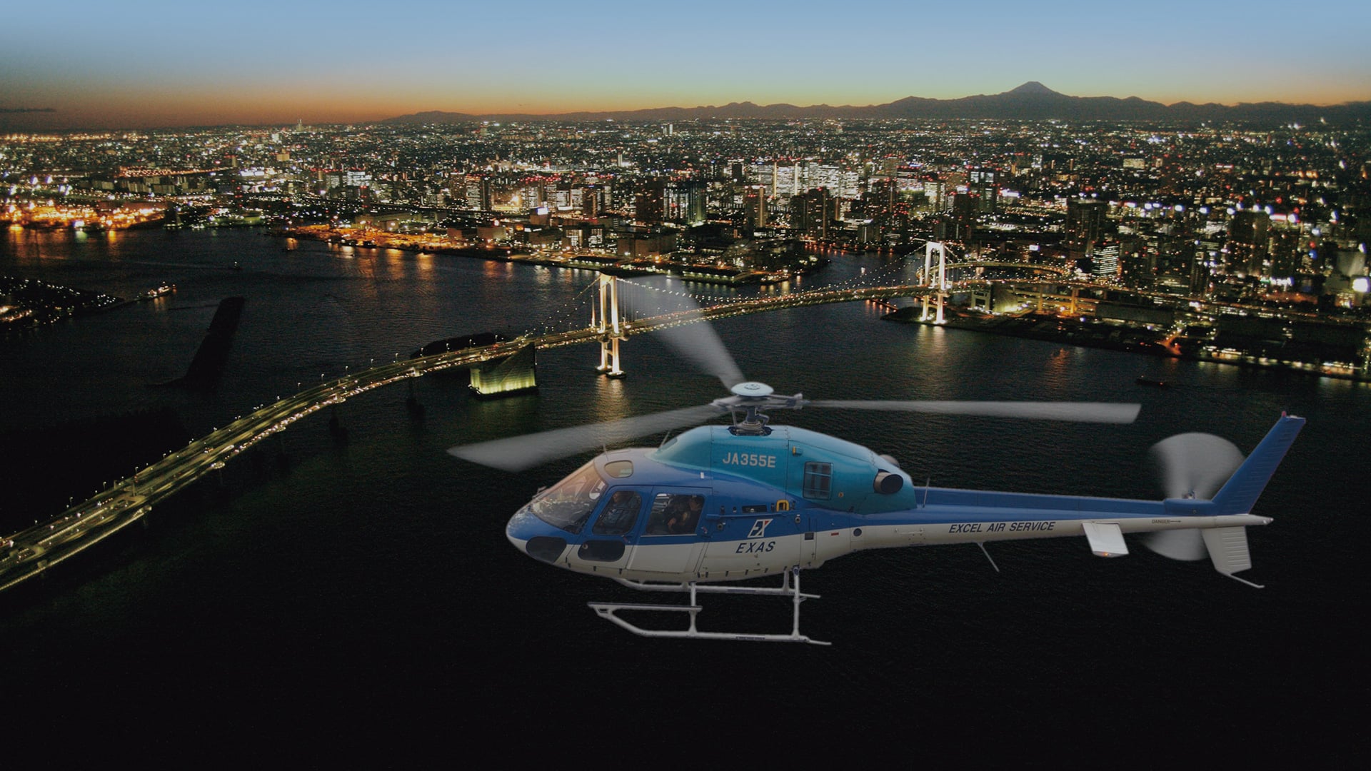 Blue and white helicopter saying: JA355E, EXASA,EXCEL AIR SERVICE flying through Tokyo.