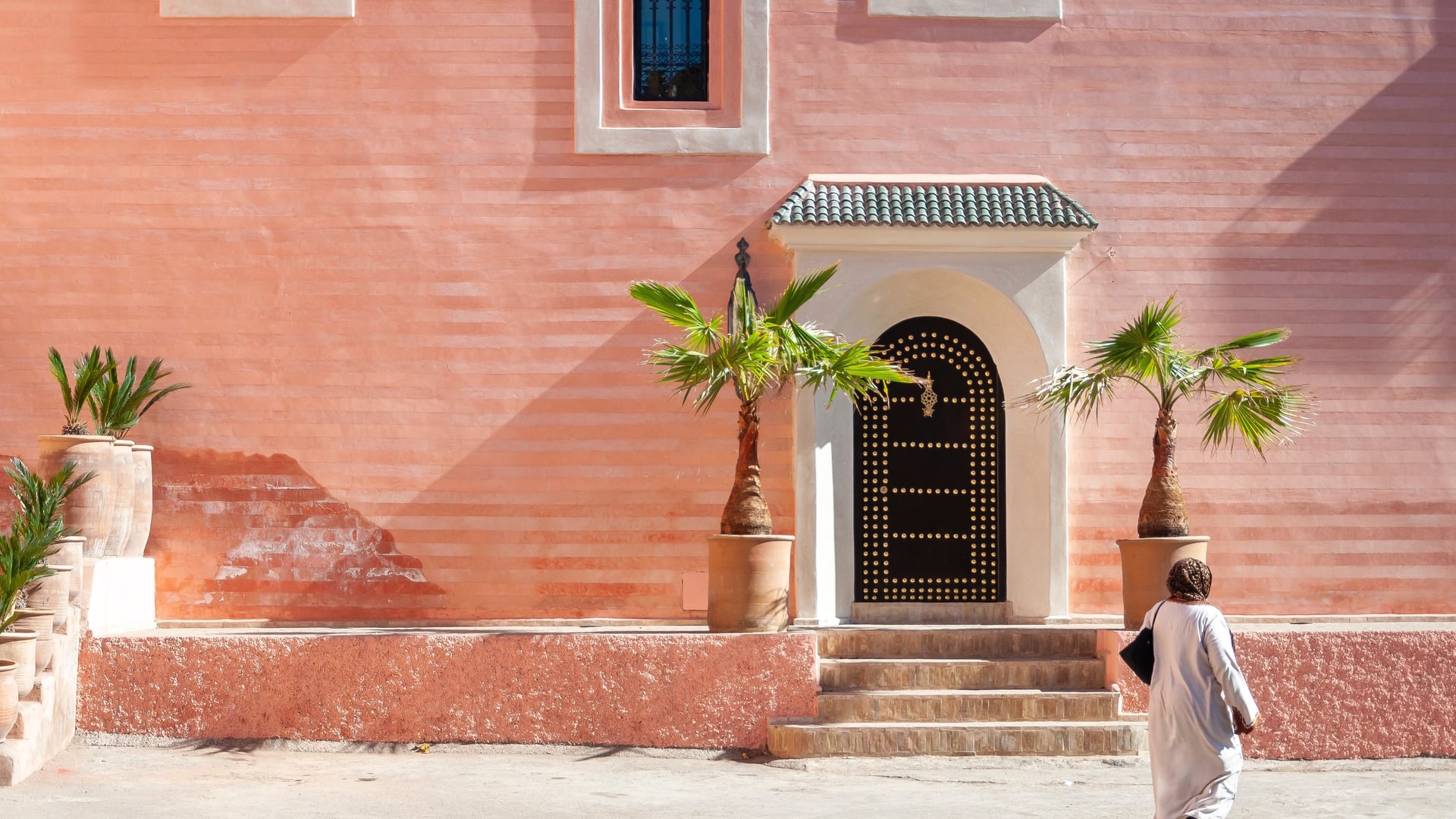 Pink building with hvite stripes, A black and gold door with two plants infront and a woman walking.