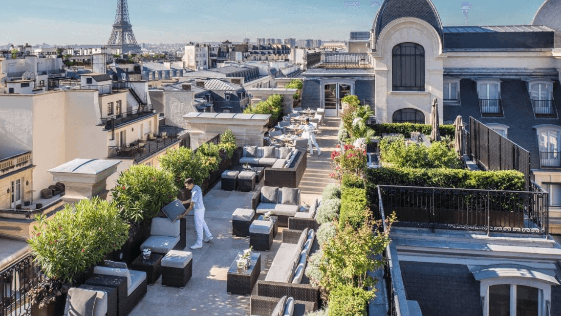 Two hotel workers in white surrounded by beautiful Paris view, plants, grey furniture at Le Rooftop - Hotel Peninsuala Paris.