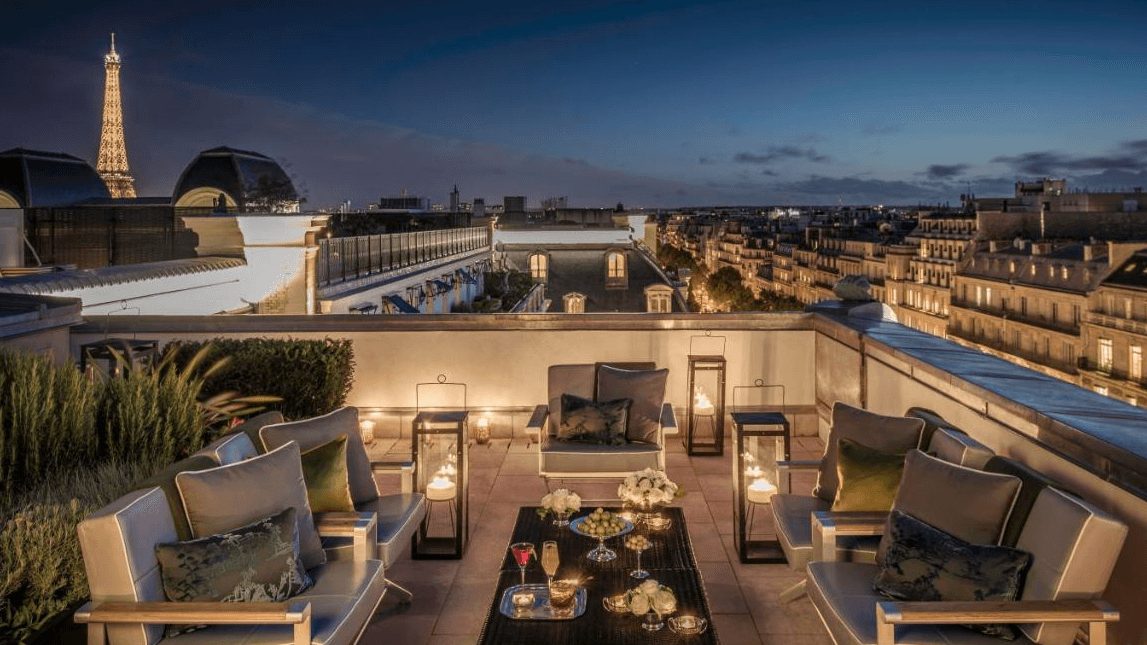 Rooftop bar in Paris with view of the Eiffel Tower, olives, and drinks at the table surrounded by plants, grey furniture and tender lightning at Le Rooftop - Hotel Peninsuala Paris.