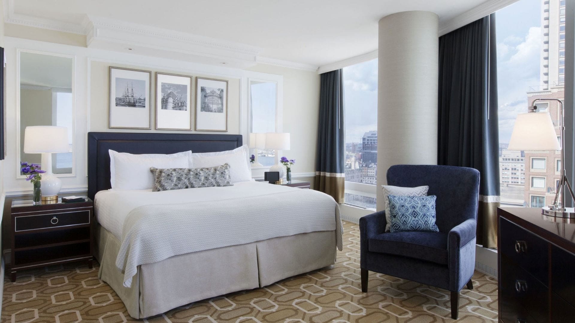 A hotel suite with a king sized bed with three paintings above. A blue chair and blue curtains.
