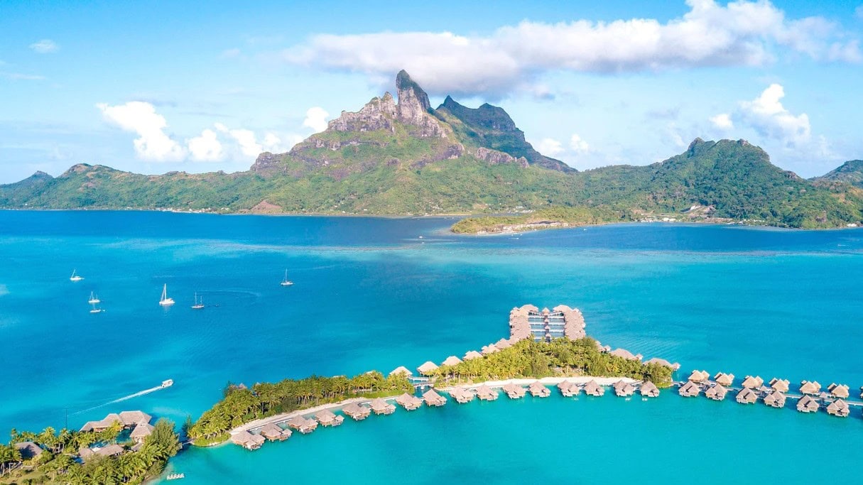 Bird view of The St Regis Bora Bora Resort with clear water, nature and mountains not far away, petite island with cabins and nature, and sailing boats in white passing by.
