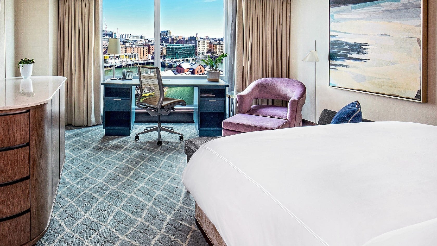 Hotel room with a bed, a blue desk, a pink chair and windows with view over the city. 